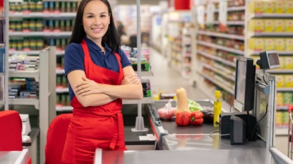 Attractive Asian woman working in supermarket standing at cash desk with arms crossed looking at camera smiling